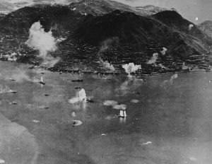 Aerial photo of an island with an urban area along its shore and a steep mountain in the center. Many ships are next to the island, and plumes of water are erupting near some of them.