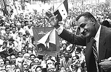 A black and white image of a man at left waving to a crowd behind him in the rest of the image that is waving Egyptian flags and holding a picture of him