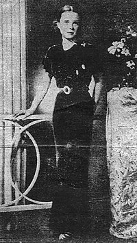 A black and white photograph of a young woman wearing a floor-length black dress with short sleeves, her right hand on a neighboring table and her left hand behind her as she faces the camera. In the middle of the image a paper crease is visible.