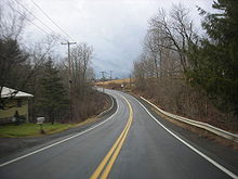 Ground-level view of a two-lane road passing through a rural area, with only one home visible on the left-hand side. Wooded terrain opens to a field as the highway curves towards the left.