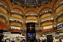 Farbfotografie einer großen Halle mit vielen Geschäften auf vier Stockwerken. Jedes Stockwerk hat mehrere runde Balkone, die rot beleuchtet sind. Das Obergeschoss hat gold-rot verzierte Rundbögen mit Wappen dazwischen und ein Kuppeldach aus Kirchenglas. Unten sind Reklametafeln und Stände von Chanel, Yves Saint Laurent und Idylle.