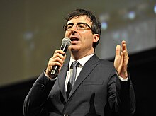 Oliver, wearing a black suit, speaking into a microphone and gesturing with the other hand