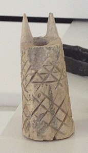 narrowish thick-walled hollow cylinder in silvery metal, tapering slightly upwards, with two triangular spikes on the top. Incised with a lattice pattern, with horizontal lines added to the top half.