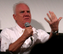 A light-skinned, white-haired elderly man talks with a microphone and gestures with his free hand.