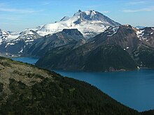 A prominent, rocky mountain with heavily glaciated lower slopes rising over mountainous terrain and an alpine lake.