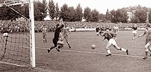 A black and white photo showing a football player kicking the ball towards the goal, while the opponent goalkeeper is trying to make a save.