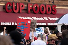 A brick storefront with the words "CUP FOODS" in red. A crowd, some with signs including a "Black Lives Matter" sign, stand before it.