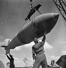 Black and white photo of a large bomb being hoisted. Two men wearing military uniforms are standing below the bomb, and steadying it with their hands.