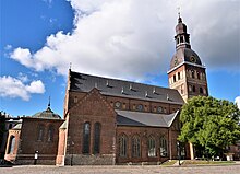 Seitliche Farbfotografie einer braunen Kirche mit einer Turmhaube an der rechten Seite, an der zwei Uhren zu sehen sind. Kleine Anbauten befinden sich um das Kirchenschiff, die lange Rundbogenfenster haben.