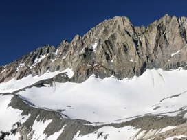 Middle Palisade presents as a long, fluted ridgeline of gray-to-brown rock above a glacier and talus moraine covered in snowfields against a blue sky