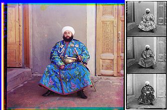 A large color photograph abutting (to its right) a column of three stacked black-and-white versions of the same picture. Each of the three smaller black-and-white photos are slightly different, due to the effect of the color filter used. Each of the four photographs differs only in color and depict a turbaned and bearded man, sitting in the corner an empty room, with an open door to his right and a closed door to his left. The man is wearing an ornate full-length blue robe trimmed with a checkered red-and-black ribbon. The blue fabric is festooned with depictions of stems of white, purple, and blue flowers. He wears an ornate gold belt, and in his left hand, he holds a gold sword and scabbard. Under his right shoulder strap is a white aiguillette; attached to his robe across his upper chest are four multi-pointed badges of various shapes, perhaps military or royal decorations.