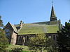A church see from the side with a gabled extension to the left and a small spire on the right; trees and a grassy bank in the foreground