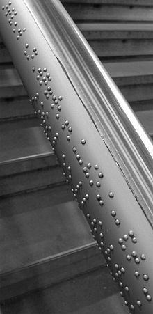 Etwa 20 cm langer Abschnitt eines Handlaufes aus grauem Stahl, im Hintergrund ist die Treppe erkennbar, an der er hinaufführt. Entlang der Seite des Stahlrohres sind mit kleinen Halbkügelchen Schriftzeichen in Braille aufgebracht.