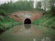 Red brick tunnel entrance through which light can just be seen at the far end. On either side are grassy banks down to the water.