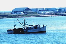 Photograph of a medium-sized fishing boat from a slight distance, as it lowers a clam dredge into the water. The photograph is taken from a perspective that shows the shore behind the boat.