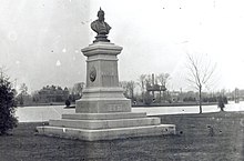 A bust of Kaiser Wilhelm I of Germany stands atop a pedestal along the shore of a small lake.