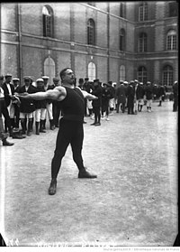 Black and white photo of man in black gymn gear holding arms outstretched, with multiple people dressed in black in the background