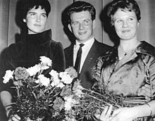 A man in a suit stands in the middle of two formally dressed women, each holding a flower bouquet.