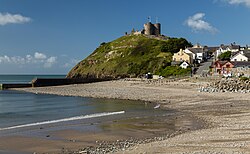 In the foreground a curved, gravelly beach. Behind it a hill rises, with a collection of houses on either side of a road on the right, and the castle rising above. It has two prominent gatehouse towers, but is otherwise low walls.