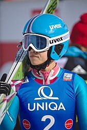 A professional skier wearing ski goggles. The skier is also wearing a blue helmet and holding skis over her shoulder.