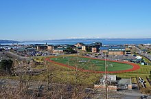 A military base with a baseball field and several buildings against a backdrop with a large body of water.