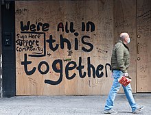 A photo of a boarded-up business in New York during the COVID-19 pandemic, featuring a masked bald man in jeans and a coat passing by; graffiti text on the boarding reads "We're ALL in this together"