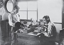 Two men working at a table with various electronic devices. One is standing on the left side of the table. The other, seated on the right side of the table, is wearing headphones and testing a crystal radio.
