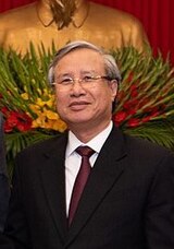 Trần Quốc Vượng seen smiling while standing in front of a bust to Hồ Chí Minh. He is wearing a black suit, a dark red tie, a white shirt and glasses.