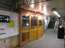 A disused oak ticket booth on the southbound platform. The booth is separated into panels that are slightly angled away from each other. Above the panels are windows, some with brass scrollwork screens.