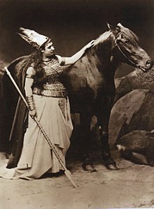 Das Schwarz-Weiß-Foto zeigt eine stehende Frau mit Brustpanzer, hohem Helm oder Kopfschmuck, Armringen und einem langen weißen Rock; sie hält in der rechten Hand einen auf den Boden gerichteten Speer und streichelt mit der linken Hand ein Pferd
