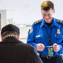 A TSA officer, smiling, looks down at a passenger's identification. The passenger is in the foreground, facing away from the camera, and is dressed in black.