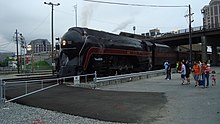 People standing next to a black streamlined steam locomotive in the foreground. The railroad signals, road bridge, and buildings are located next to the locomotive in the background.