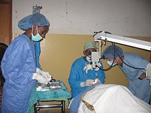 Surgeon using surgical microscope to operate while theatre staff attend.