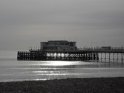 The end of a pier under cloudy skies, illuminated by sunlight reflecting off a calm sea. There is a small stretch of rocky beach in the foreground.