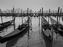 Black-and-white photo on a gray day. In the foreground, four long, narrow boats float side-by-side, left to right, each loosely moored to one of the four tall poles standing in the water (two to each side). Some 30 meters away, in the background, a further row of 15 or 16 gondolas can be seen similarly moored near a railed walkway on the far side. Buildings of Venice appear as distant shadows in the mist.