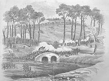 A 19th century engraving of a landscape in which a military action is taking place. Two opposing forces of soldiers battle over a stone bridge. Two flags are carried by soldiers at the center of the bridge. Troops approach from a high hill in the background and white smoke rises on the hillside.
