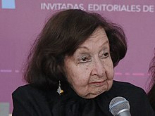 Amparo Dávila sitzt vor einem Tisch, wo ihre Name steht und ihrem Buch Árboles petrificados. Eine Frau hält das Mikrofon, damit sie sprechen kann, weil sie bereits sehr alt ist.