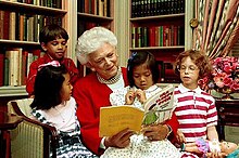 This is a photo of Former First Lady, Barbara Bush, on a couch in the White House Library. She is surrounded by a handful of children crowing her while she reads a book.
