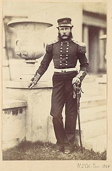 A bearded man in Victorian military uniform poses in front of an ornamental stone urn