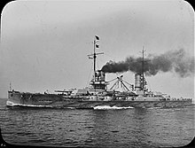 A large gray battleship steams ahead. Dark black smoke pours from its two funnels.