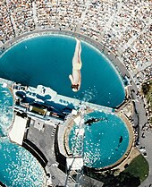 Das Foto zeigt einen Springer in der Luft über einem Wasserbecken und einer Zuschauertribüne.