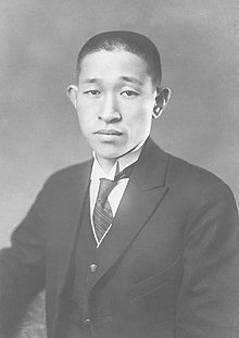 Black and white portrait. Head shot of Matsushita in front of a dark background, wearing a dark suit jacket with a light wing-collared shirt, dark vest, and dark striped necktie.