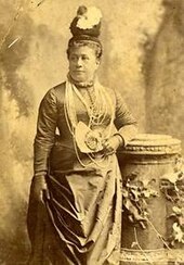 Photograph of Queen Kapiʻolani standing looking to her left. She is wearing a hat and a necklace of several strands of Ni'ihau shells.