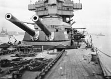 A twin-barrelled gun turret on a large warship, photographed by someone standing near the bow. Anchor chains are in the foreground, and the superstructure of the ship can be seen behind the turret.