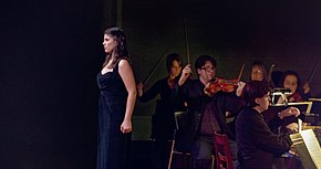 Frau mit langen dunklen Haaren in dunkelblauem Kleid steht nach rechts gewandt vor schwarzem Hintergrund, hinter ihr halten Musiker mit Streichinstrumenten ihre Streichbögen hoch, ganz links sitzt ein Pianist.