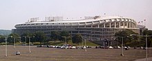 A large circular stadium with a curving overhang behind a mostly empty parking lot.