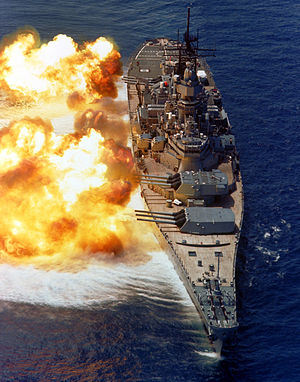 USS Iowa in her post 1980s rebuild. The battleship is pointed toward the viewer, with her 9 gun barrels pointed starboard for a gunnery exercise. Fire can be seen erupting from the gun barrels, and a concussive effect is visible on the water.