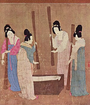 A group of women wearing high-waisted skirts, wrap-front tops and large hair buns use wooden rods to prepare a length of white silk.