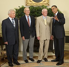 The astronauts are all elderly but standing straight. Aldrin wears a dark suit, Collins a dark sport coat and gray pants, and Armstrong a beige suit. The President is at the right. He wears a dark suit. He has medium-dark skin and is talking to Armstrong and raising his left hand. Armstrong is smiling.