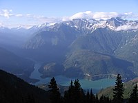 Landscape of tall forested mountains with snow caps above the treeline and blue lake below
